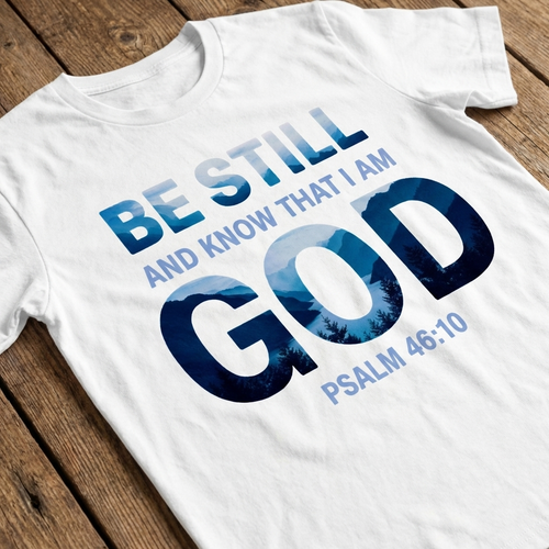 White t-shirt with 'Be still and know that I am God' text on a wooden surface