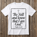 White t-shirt with 'Be Still and know that I am God' text on a wooden background