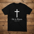 Black t-shirt with a white cross and 'He is Risen' text on a wooden surface