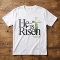 White t-shirt with 'He is Risen' text and cross design on a wooden surface