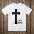 White t-shirt with a black cross and 'He is Risen' text on a wooden background