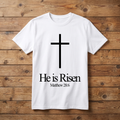 White t-shirt with a black cross and 'He is Risen' text on a wooden background
